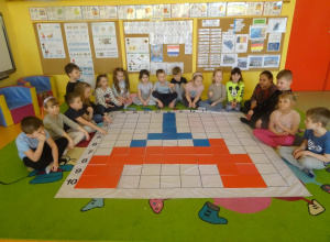 Grupa dzieci siedzi wokół maty na której kolorowe płytki ułożone są na kształt wieży.